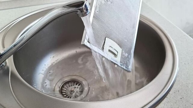 Running water from a kitchen faucet rinses a metallic mesh filter in a stainless steel sink, demonstrating household cleaning, hygiene, and appliance maintenance practices