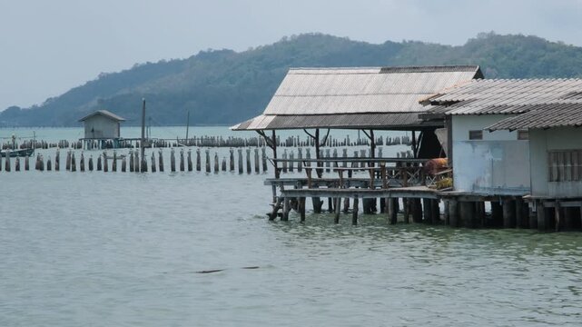 Wooden stilt buildings extending over calm sea water with a covered dock structure and lush green hills visible in the background in Thailand