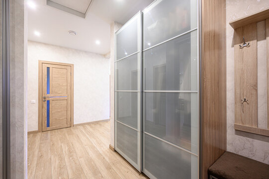 A modern hallway with light wooden flooring, frosted glass sliding wardrobe doors, wooden door with blue accents, built-in hooks, bench, light-colored walls, recessed ceiling lights.