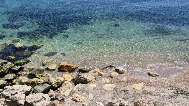 Crystal clear turquoise water gently laps against a rocky shoreline. Sun-drenched stones and golden sand sit beneath the transparent waves, transitioning into a deep blue sea under bright light.