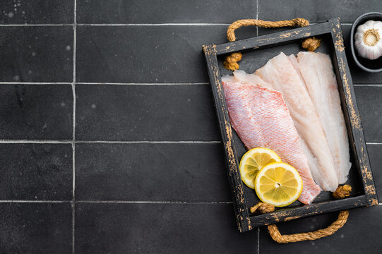 Raw redfish fillets on rustic wooden tray with lemon slices. Fresh seafood ingredient on dark background with copy space.