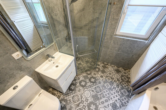 A modern bathroom with a glass shower cabin, white sink with cabinet, toilet, patterned floor tiles, gray marble-like walls, radiator, two windows with blinds, clean minimalist design.