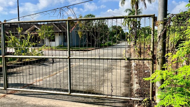 Old metal gate blocking a quiet residential street with overgrown vegetation under a bright blue sky in a tropical neighborhood.