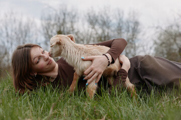 Naklejka premium Relaxed woman outdoors, cuddling a curious goat kid in a sunny grassy field, joyful moment, soft breeze, natural colors, serene rural scene, warm connection between human and animal