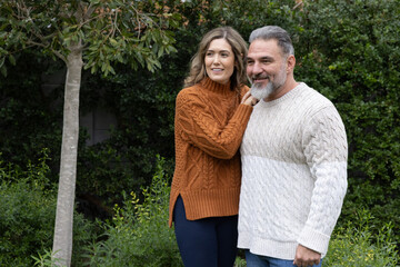 Fototapeta premium Couple standing in private garden wearing cable-knit sweaters by thin-trunked tree and hedges