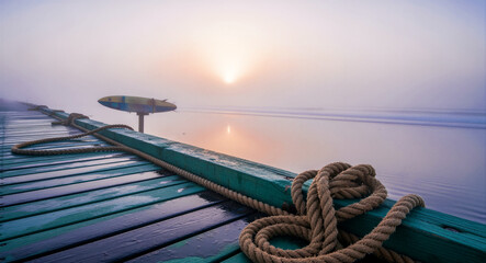 Naklejka premium Nautical ropes resting on a wooden pier overlooking a misty beach at sunrise. Soft morning light illuminates the fog over calm ocean waves. Coastal serenity concept, ideal for travel marketing.