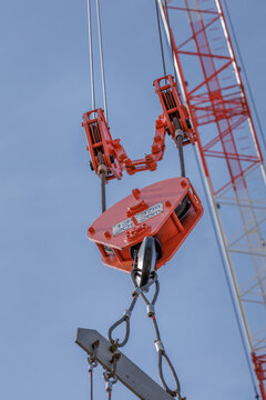 Close-up portrait red crane hook block and pulley rigging gripping steel cables attached to lifting beam, set against clear blue sky, with part of crane&rsquo;s lattice boom rising in bokeh background.