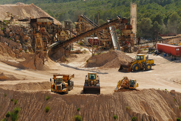 Heavy machinery working at limestone quarry with conveyor system and crushing plant in background. Industrial mining site with excavator, loader and dump truck for aggregate production. © Ekaterina