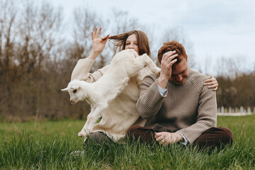 Naklejka premium A couple sits on lush grass outdoors with a playful goat kid nearby, capturing a warm lifestyle moment of joy, bonding, and simple countryside happiness.