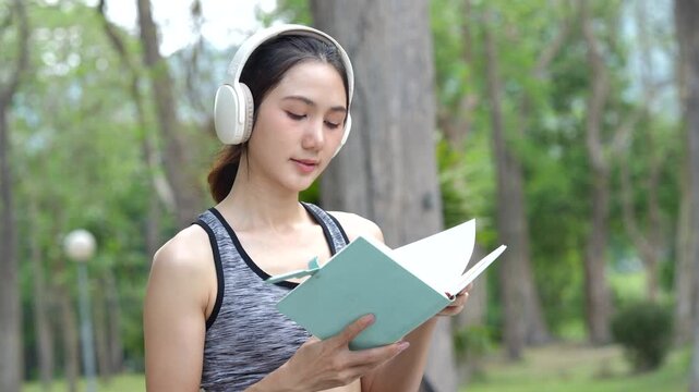 A young woman wearing headphones reads a notebook in a park, enjoying music and learning outdoors after exercise in a calm natural setting.