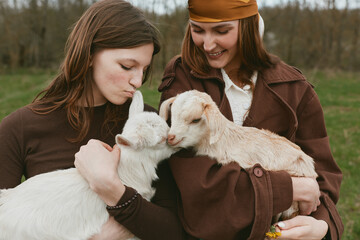 Naklejka premium Goat, kid, lifestyle, outdoor scene with two women embracing gentle animals in a sunlit field, candid interaction, warm textures, casual outfits, rustic backdrop, wholesome companionship, nature