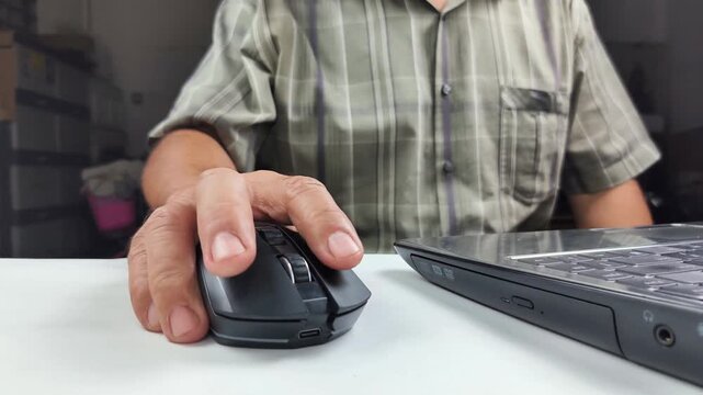 Close-up of a man's hands using a wireless computer mouse on a desk, spinning the wheel and clicking buttons while inputting data via a laptop keyboard.