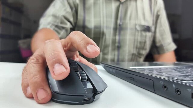 Close-up view of a man's hand using a wireless computer mouse on a desk, scrolling the wheel and clicking the buttons while browsing the internet.