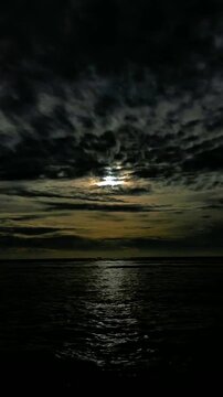A sliver of moonlight breaks through heavy clouds, reflecting on the dark, rippling water below, creating a stark and dramatic seascape at night.