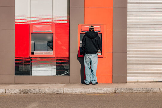 Young man in black hoodie and baggy jeans withdrawing cash at a red ATM on a sunny urban street