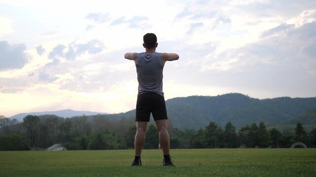 Cinematic back view of a male athlete performing a standing full body stretch in a park, scenic mountain landscape at sunset background, wellness and healthy lifestyle concept