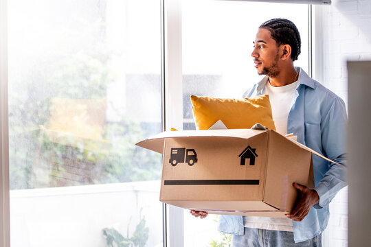 Young man holding a cardboard box with household belongings, moving into a bright new apartment, concept of home, independence and a new beginning