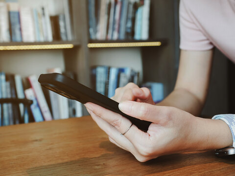 Young woman holding smartphone over wooden table with bookshelf background showing relaxed focus and warm lighting