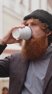 Joyful young man enjoying morning coffee or tea hot drink outdoors. Relaxing, taking a break. Redhead bearded boy walking in urban city center street, drinking coffee to go. Town lifestyles outside