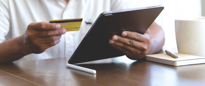 Person holding credit card and using tablet for online shopping at home, with coffee and notebook on wooden desk, ideal for e-commerce, digital payment, fintech, and modern lifestyle concepts