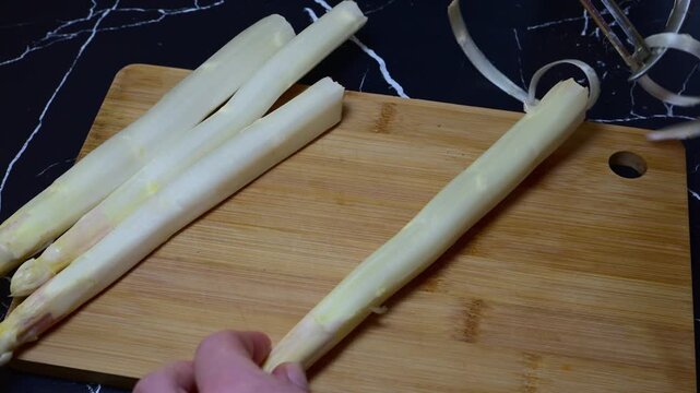 hand using a silver peeler to remove the tough outer skin from a fresh white asparagus stalk on a wooden cutting board