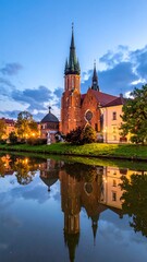 Fototapeta premium Illuminated church tower and building reflect in calm water at dusk, with trees and a cloudy sky