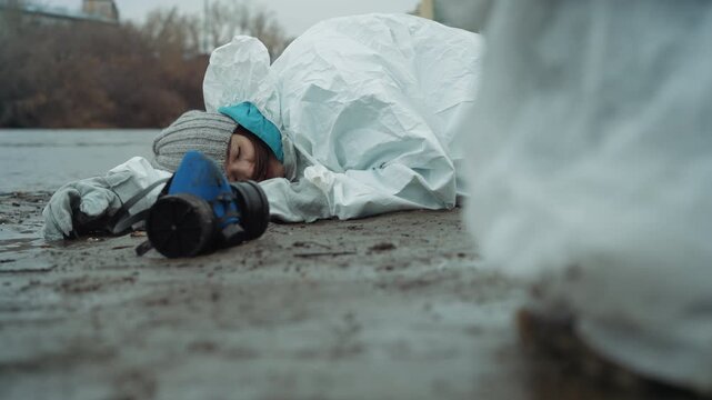 Young survivor lying by icy riverbank, responder kneels nearby in protective suit, gasmask on muddy ground, inspection and retrieval underway, decontamination protocol in motion, cold gray sky