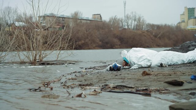 Abandoned protective suit on riverbank, tattered white coverall lies in mud near gray water and leafless shrubs, distant industrial buildings on horizon, late winter frost, scattered debris