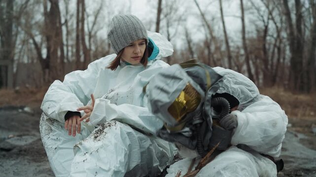 White scientist and child in hazmat sit on frozen roadside amid leafless trees, sharing tired gaze over muddy boots and broken shovel abandoned town atmosphere, cold breath, protective gear smeared