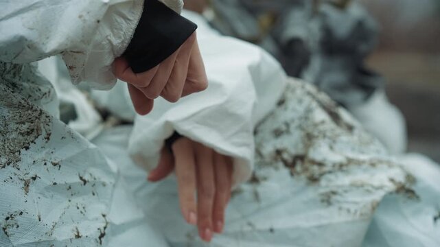 Cold hands in protective suit, mudstained sleeves and trembling fingers, closeup of child survivor resting on torn fabric, abandoned urban background, frostdusted textures, bleak atmosphere of crisis