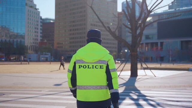 Police officer in bright neon yellow high visibility jacket patrols urban plaza on sunny winter day with modern office buildings, bare trees casting long shadows, and pedestrians in background.