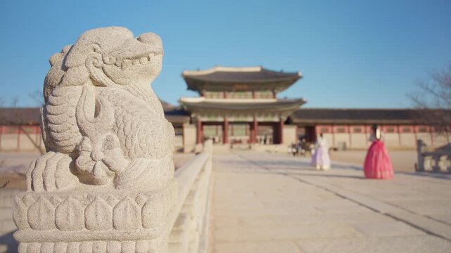 Ancient stone guardian statue on bridge railing at Gyeongbokgung Palace in Seoul South Korea with women wearing traditional hanbok dresses walking toward historic gate under clear blue sky.