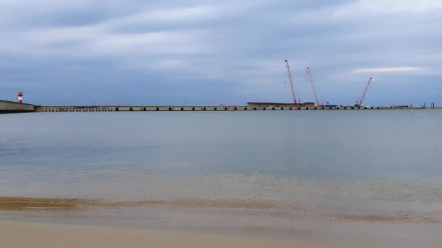 Empty seaport harbor on Baltic sea shore with calm coastal surf water waves rolling on sand beach and port quayside on background under cloudy sky at overcast day. No people serene seaside landscape.