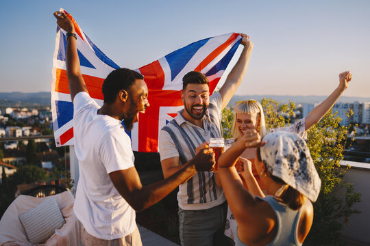 Young friends cheering and toasting beer on a rooftop while holding a british flag