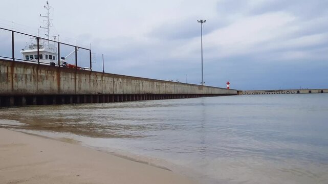 Low angle view quay wall at empty seaport harbor on Baltic sea shore with calm coastal surf waves rolling on sand beach under cloudy sky at overcast day. No people port quayside seaside landscape.