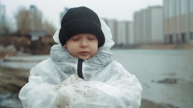 Caucasian child in protective suit by icy shore, black beanie, distant highrise skyline, abandoned waterfront, solemn expression, hands clasped, frost on sleeves, cold breath, implied nuclear fallout,