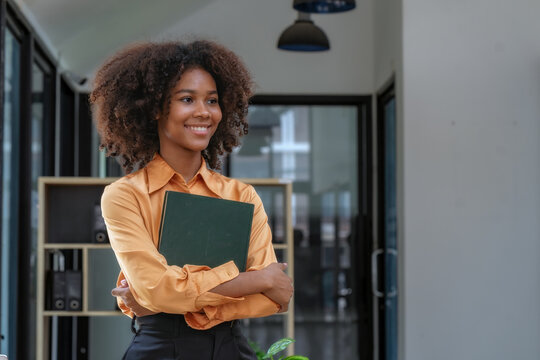 Smiling African American young woman holding notebook and looking away in modern office.