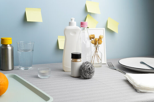 Modern still life of household items. Cleaning supplies, dishes, food on a gray tablecloth against a blue wall with yellow sticky notes. Soft lighting creates a clean, conceptual aesthetic