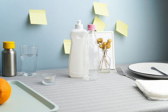 A contemporary still life of everyday household items on a table. The scene includes plastic bottles, fruit, and tableware against a blue wall with yellow sticky notes. Soft light and pastel colors