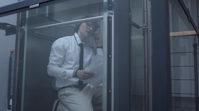 Office worker talks with clients on the phone in a glass elevator while reviewing papers capturing business communication focus responsibility and a busy professional routine