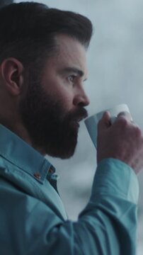 Interior scene captures contemplative man in teal shirt savoring coffee near frosty window with snowy outlook