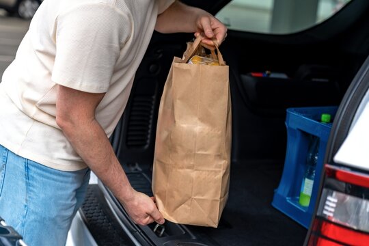 Individual packing supplies into vehicle. Person organizing shopping items into car trunk. Man dressed casually placing shopping bags and coolers into car in suburban neighborhood