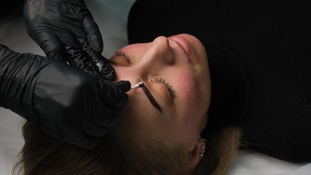 Master in black gloves correcting the shape of a client's eyebrows after a cosmetic procedure