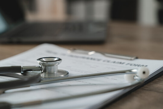 Stethoscope placed on medical claim form on clipboard with blurred laptop background, representing health insurance documentation, medical verification process, patient record review, and healthcare c
