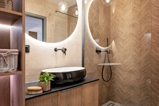 Interior of a modern bathroom with black and white modern wash basin on wooden cabinet and design tile in herringbone on the wall. Apartment with wood, glass and stylish decor.