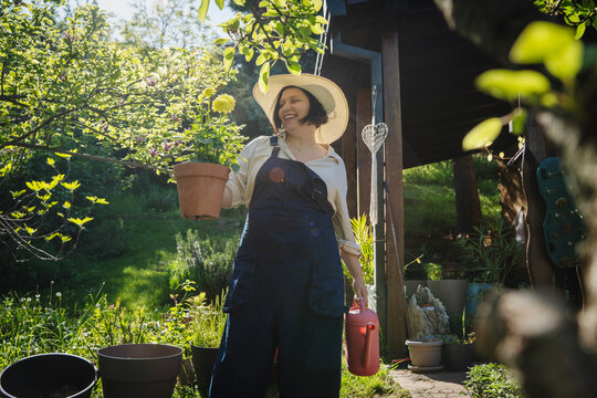 Happy caucasian woman in hat holding flower pot on house terrace in summer sun