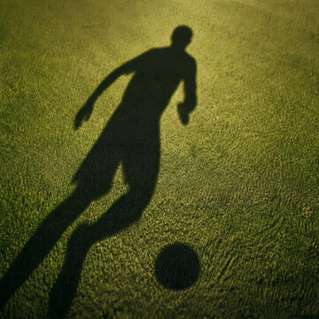 dynamic soccer player shadow dribbling on green grass pitch long late afternoon sunlight ball shadow separate abstract athlete motion no visible player aerial top down view striking contrast sports co