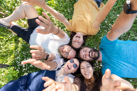 Five happy adult friends lying in a circle head to head on grass waving at camera
