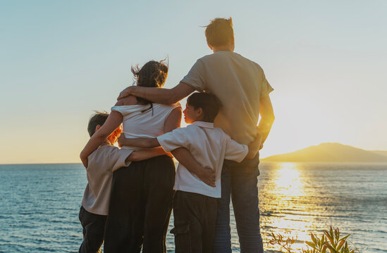 Young family with two sons hugging and watching sunset at the sea, travel and togetherness concept