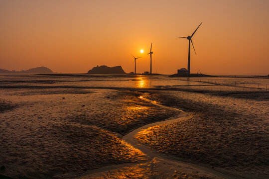 Dramatic sunset at Tando Port in Ansan, South Korea, featuring wind turbines and the Nue Island Lighthouse Observatory with golden light reflecting on the winding mudflat water trail.